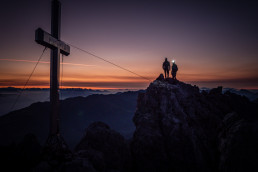 Bergsteiger mit Gipfelkreuz vor dramatischer Kulisse im Sonnenuntergang